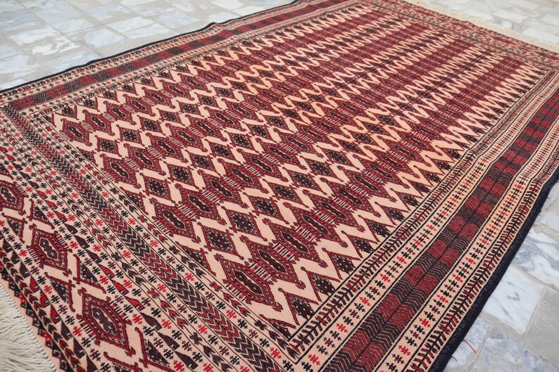 May include: A traditional rug with a geometric pattern in shades of red, black, and cream. The rug has a fringed border.