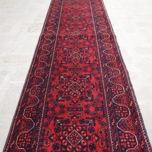May include: A long, rectangular red rug with intricate geometric patterns. The rug features a central field of red with navy blue and orange accents. The border is navy blue with a repeating white and orange design.
