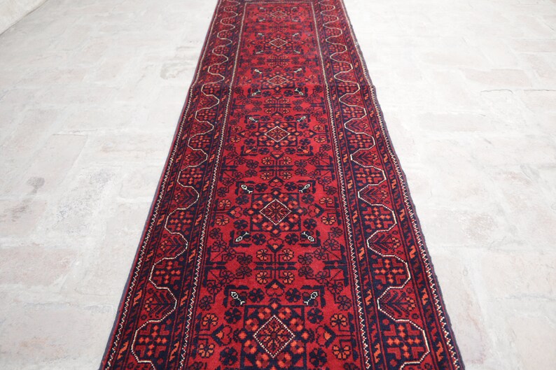 May include: A long, rectangular red rug with intricate geometric patterns. The rug features a central field of red with navy blue and orange accents. The border is navy blue with a repeating white and orange design.