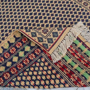 May include: A close-up of a woven rug with a geometric pattern in shades of beige, blue, red, and green. The rug has fringe on all sides.
