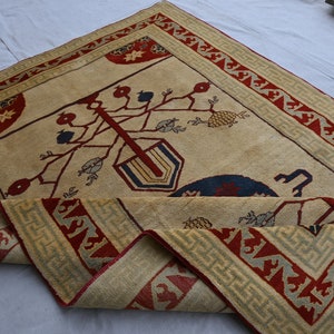 May include: A beige rug with a red and green border. The center of the rug features a stylized tree with red, blue, and yellow details. The rug is folded in half.