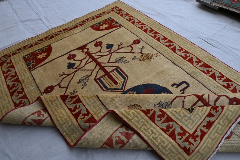 May include: A beige rug with a red and green border. The center of the rug features a stylized tree with red, blue, and yellow details. The rug is folded in half.