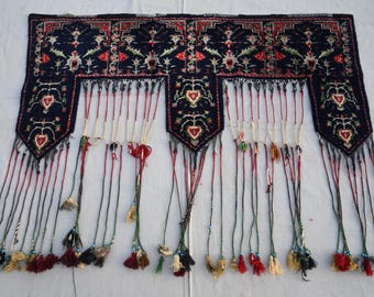 Op de afbeelding: Een donkerblauw, rechthoekig textiel met ingewikkelde bloemen- en geometrische patronen in rood, wit en groen. Het textiel heeft drie hangersecties met hartvormige ontwerpen en lange, bungelende kwasten in verschillende kleuren.