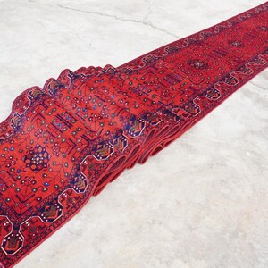 May include: A red runner rug with a traditional Persian design featuring intricate floral and geometric patterns in shades of blue, red, and white. The rug is folded in half and has a fringe on the ends.