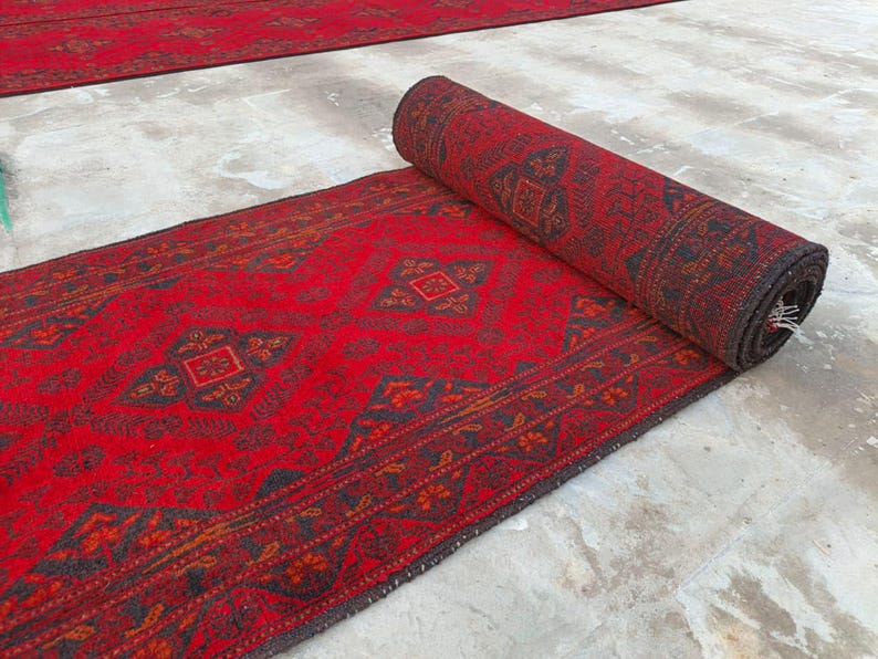K&ouml;nnte beinhalten: Eine aufgerollte rote Teppichrolle mit detailliertem geometrischem Muster. Der Teppich hat einen dunklen Rand und komplizierte Designs in Rot-, Braun- und Schwarzt&ouml;nen. Der Teppich ist teilweise ausgerollt.