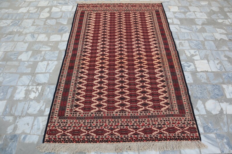 May include: A traditional rug with a red, black and white geometric pattern. The rug has a fringe border and is made of wool.