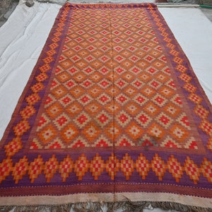 Op de afbeelding: Een groot, rechthoekig tapijt met een geometrisch patroon in tinten oranje, rood en paars. Het tapijt heeft een franje rand.