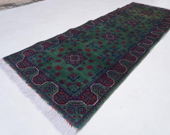 May include: A rectangular, green rug with a detailed pattern in red and navy blue. The rug has a central field of green with repeating floral motifs and a decorative border. White fringe edges the rug.