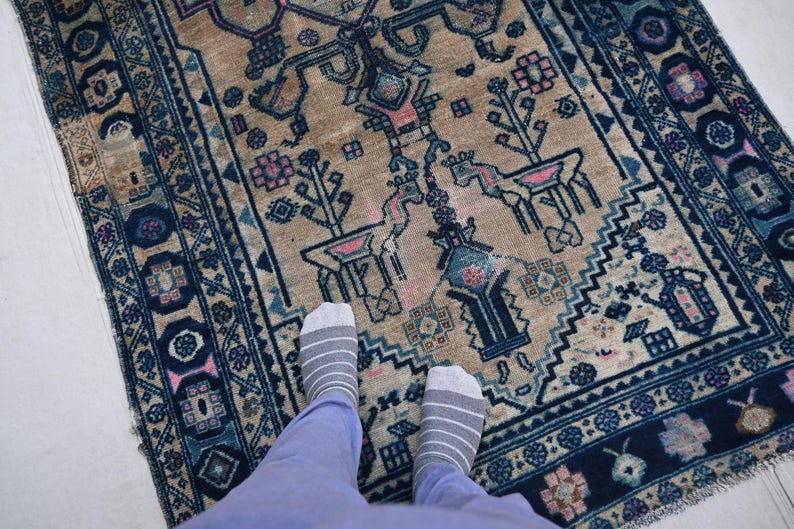 Peut inclure: Un tapis beige antique avec une bordure bleu marine d&eacute;taill&eacute;e et un motif central. Le tapis pr&eacute;sente des motifs floraux et g&eacute;om&eacute;triques, avec des accents roses et bleus. Le tapis est rectangulaire et semble fait &agrave; la main.