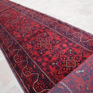 May include: A long, rectangular red rug with a detailed geometric pattern in navy blue and accents of gold. The rug is partially rolled up, revealing the underside with a similar design. The rug is likely made of wool.