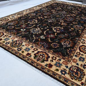 May include: A dark brown rug with a cream-coloured border. The rug has a floral pattern with intricate details. The rug is fringed on all sides.