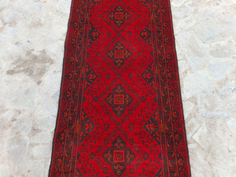 K&ouml;nnte beinhalten: Ein rechteckiger roter Teppich mit einem sich wiederholenden geometrischen Muster in Schwarz und Braun. Der Teppich hat ein zentrales Design aus Rauten und einen Rand mit floralen Motiven. Der Teppich liegt auf einer hellen, strukturierten Oberfl&auml;che.