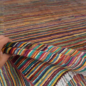 May include: A woven rug featuring a dense pattern of thin, horizontal stripes in vibrant shades of red, yellow, blue, green, and purple. The textile has a textured surface and is finished with short, white fringe along the edge of the fabric.