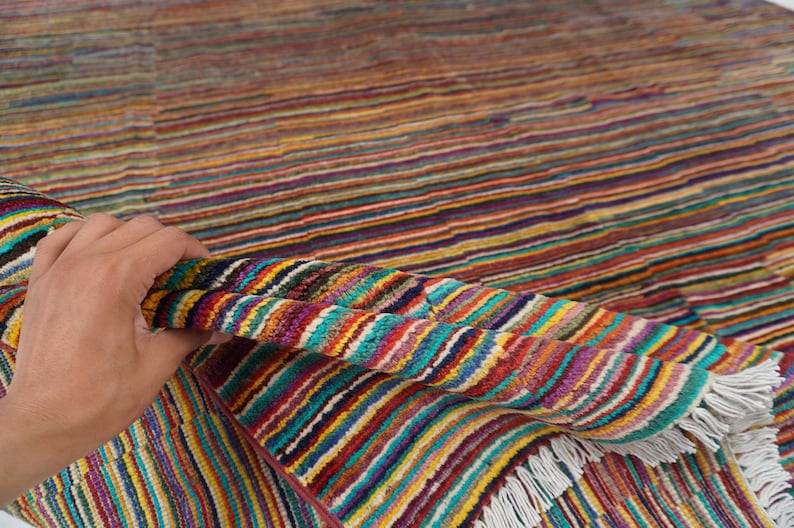 May include: A woven rug featuring a dense pattern of thin, horizontal stripes in vibrant shades of red, yellow, blue, green, and purple. The textile has a textured surface and is finished with short, white fringe along the edge of the fabric.