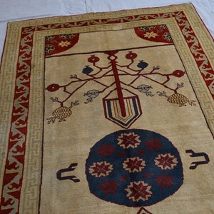 May include: A beige rug with a red and green border. The center of the rug features a stylized tree with red and blue fruit, and a blue circle with red starbursts.