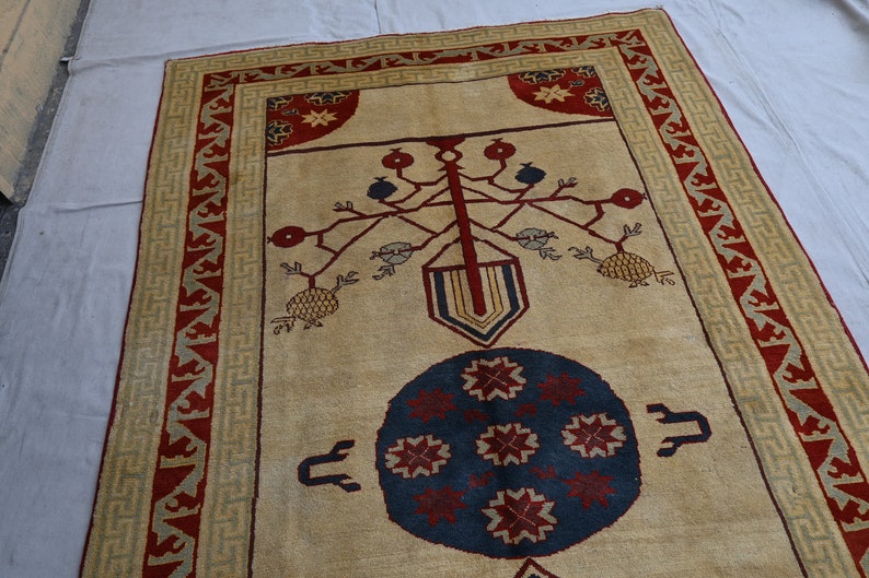 May include: A beige rug with a red and green border. The center of the rug features a stylized tree with red and blue fruit, and a blue circle with red starbursts.