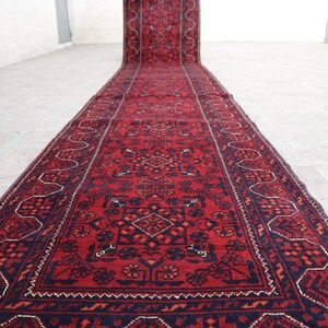 May include: A long, rectangular red rug with intricate geometric patterns in navy blue and orange. The rug is rolled out on a light-colored surface, showcasing its detailed design and rich color palette. The rug's border features a repeating pattern.