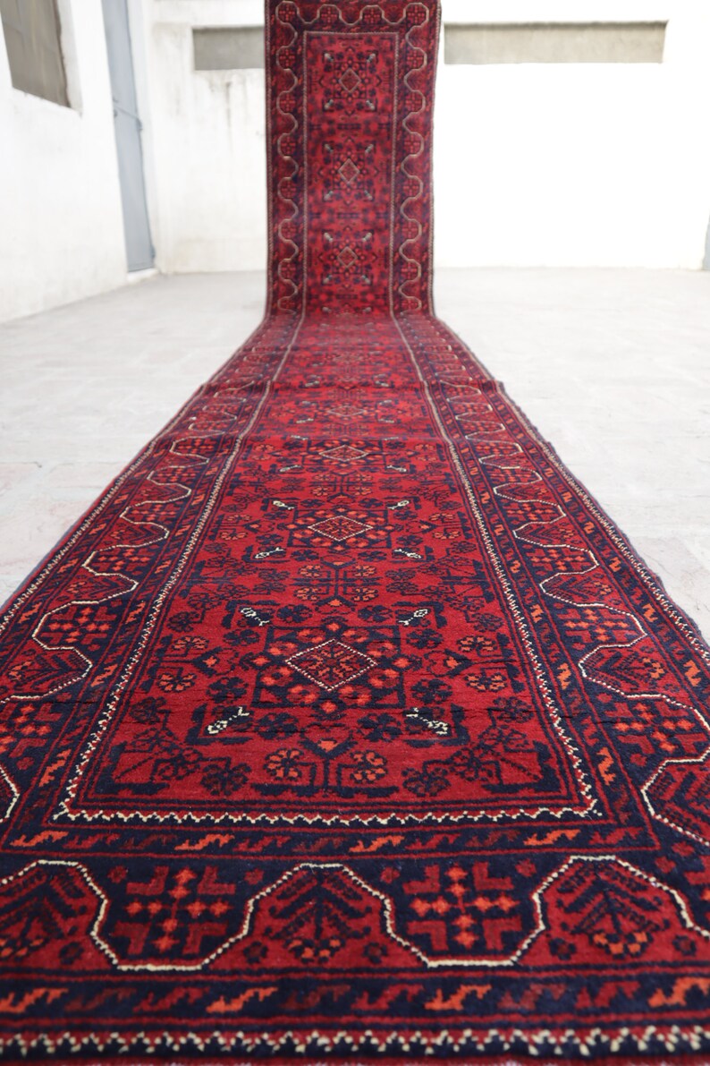 May include: A long, rectangular red rug with intricate geometric patterns in navy blue and orange. The rug is rolled out on a light-colored surface, showcasing its detailed design and rich color palette. The rug's border features a repeating pattern.