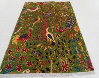 May include: A rectangular green rug with a tree and bird design. The rug features a central brown tree with birds, peacocks, and flowers in red, yellow, and blue. The rug has a white fringe.