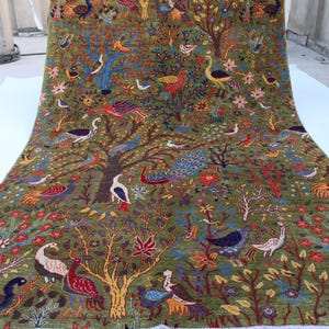 Pode incluir: Um tapete colorido com fundo verde que apresenta um padrão repetitivo de árvores estilizadas, pássaros e pavões. O tapete é tecido com detalhes intrincados e cores vibrantes.