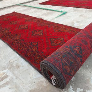 K&ouml;nnte beinhalten: Rot-schwarzer Teppich mit Muster, teilweise aufgerollt, mit einem weiteren &auml;hnlichen Teppich im Hintergrund. Der Teppich hat ein geometrisches Design und einen dunklen Rand. Der aufgerollte Teppich zeigt die Unterseite mit Fransen. Die Teppiche liegen auf einer Betonoberfl&auml;che.