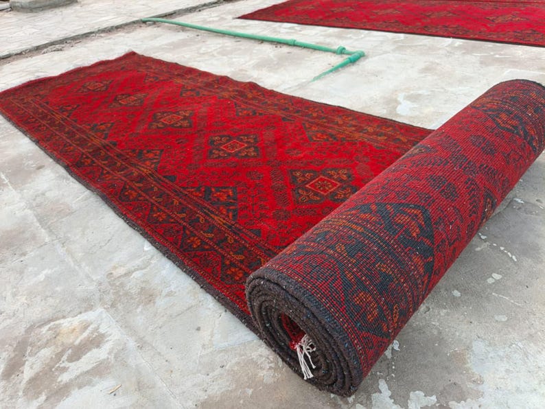 K&ouml;nnte beinhalten: Rot-schwarzer Teppich mit Muster, teilweise aufgerollt, mit einem weiteren &auml;hnlichen Teppich im Hintergrund. Der Teppich hat ein geometrisches Design und einen dunklen Rand. Der aufgerollte Teppich zeigt die Unterseite mit Fransen. Die Teppiche liegen auf einer Betonoberfl&auml;che.