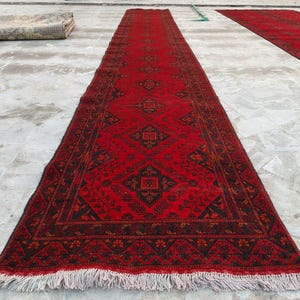 K&ouml;nnte beinhalten: Ein langer, rechteckiger roter Teppich mit einem detaillierten geometrischen Muster in Schwarz und Braun. Der Teppich hat Fransen an den Enden und wird im Freien ausgestellt. Der Teppich ist wahrscheinlich aus Wolle oder einem &auml;hnlichen Material.