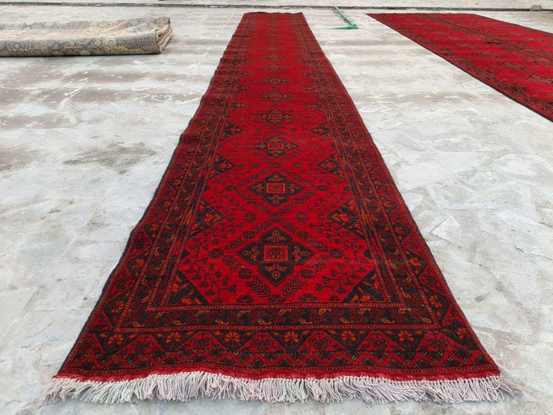 K&ouml;nnte beinhalten: Ein langer, rechteckiger roter Teppich mit einem detaillierten geometrischen Muster in Schwarz und Braun. Der Teppich hat Fransen an den Enden und wird im Freien ausgestellt. Der Teppich ist wahrscheinlich aus Wolle oder einem &auml;hnlichen Material.