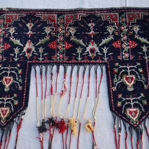 Op de afbeelding: Een donkerblauw textiel met ingewikkelde bloemen- en geometrische patronen in rood, wit en groen. Het textiel heeft een gefranjerde rand met kwasten in verschillende kleuren. Het ontwerp bevat hartvormige motieven.
