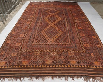 May include: A rectangular woven rug featuring a deep rust and brown color palette with intricate geometric patterns. The central field displays a series of stacked diamonds, surrounded by multiple decorative borders and finished with a fringed edge.