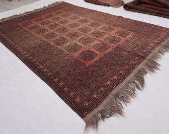 Puede incluir: Una alfombra rectangular con una paleta de colores rojo oscuro y marrón, con un campo central de motivos octogonales repetitivos. La alfombra está enmarcada por un borde ancho e intrincado con patrones florales y flecos marrón claro.