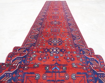 May include: A rolled-up, long, red runner rug with a detailed geometric pattern in blue, green, and white. The rug's design features floral and geometric motifs, creating a traditional aesthetic. The rug is displayed on a light-colored surface.