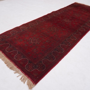 May include: A long, rectangular, red rug with a detailed pattern and fringe on both ends. The rug has a central field of red with a repeating geometric design and a border with a wavy pattern.