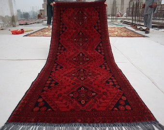 Puede incluir: Una alfombra roja y negra tejida a mano con intrincados patrones florales y un borde con flecos. La alfombra está siendo sostenida por una persona, mostrando su longitud completa.