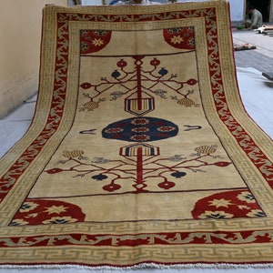 May include: A large, cream-colored rug with a red and gold border. The rug features a central medallion with a blue and red design, and two stylized trees with red and blue fruit.