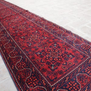 May include: A long, rectangular red rug with intricate geometric patterns in navy blue and accents of gold. The rug features a central design with repeating floral and diamond motifs, bordered by a detailed navy blue edge.