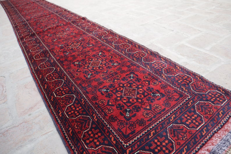 May include: A long, rectangular red rug with intricate geometric patterns in navy blue and accents of gold. The rug features a central design with repeating floral and diamond motifs, bordered by a detailed navy blue edge.