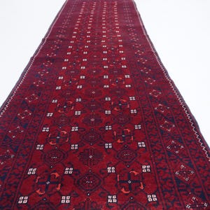 May include: A long, rectangular, red rug with a detailed geometric pattern. The rug features a central design with repeating shapes and borders in dark blue and white. The rug is likely made of woven material.