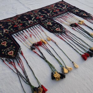Op de afbeelding: Een donkerblauw, rechthoekig textiel met ingewikkelde bloempatronen en hartvormige ontwerpen. De randen zijn versierd met lange kwasten in verschillende kleuren, waaronder rood, zwart en goud. Dit decoratieve stuk lijkt een tapijt of wandkleed te zijn.