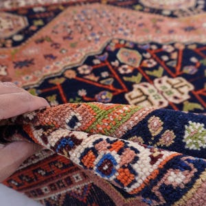 May include: A rolled-up section of a Persian rug, showcasing intricate patterns and a rich color palette. The rug features a navy blue background with floral and geometric designs in shades of peach, orange, and green. The fringe is visible at the edge.