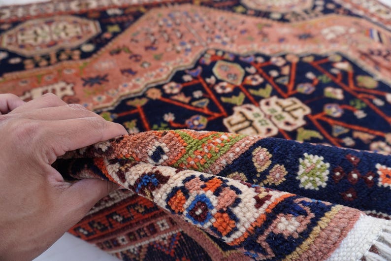 May include: A rolled-up section of a Persian rug, showcasing intricate patterns and a rich color palette. The rug features a navy blue background with floral and geometric designs in shades of peach, orange, and green. The fringe is visible at the edge.