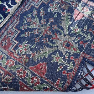Op de afbeelding: Close-up van een gedeelte van een donkerblauw tapijt met een bloemenpatroon in rood, groen en wit. Het tapijt heeft een gedetailleerde rand en franjes. Het tapijt is dubbelgevouwen, waardoor de onderkant zichtbaar is.