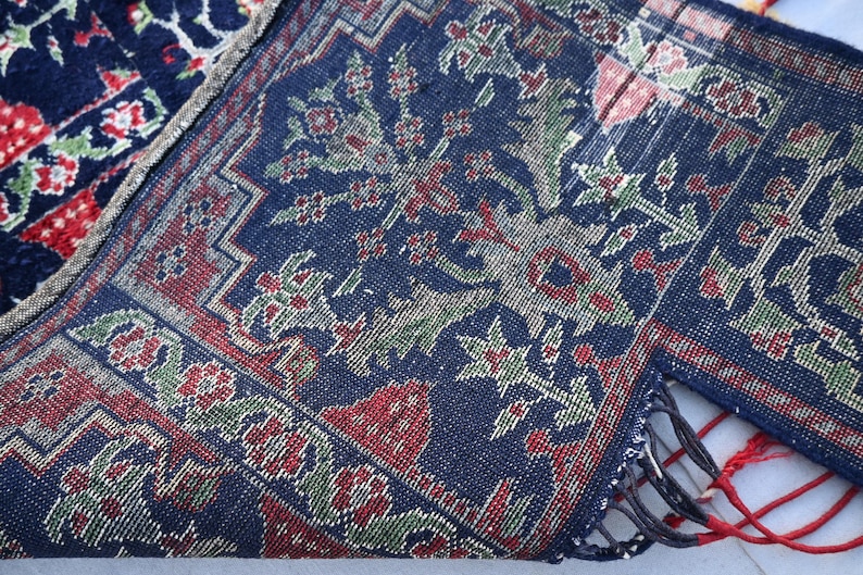 Op de afbeelding: Close-up van een gedeelte van een donkerblauw tapijt met een bloemenpatroon in rood, groen en wit. Het tapijt heeft een gedetailleerde rand en franjes. Het tapijt is dubbelgevouwen, waardoor de onderkant zichtbaar is.