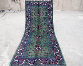 May include: A green rug with a traditional Persian design. The rug has a dark blue border with red and white accents. The center of the rug features a repeating pattern of diamonds and floral motifs.