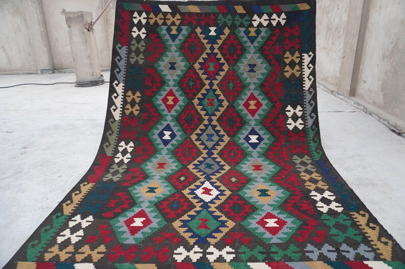 May include: A traditional hand-woven rug with a geometric pattern in shades of red, green, blue, and white on a dark brown background.