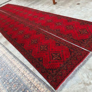 K&ouml;nnte beinhalten: Zwei lange, rechteckige rote Teppiche mit komplizierten geometrischen Mustern in Schwarz und Braun. Die Teppiche haben an den Enden Fransen und sind nebeneinander platziert. Ein kleinerer, gemusterter Teppich in Blau- und Beiget&ouml;nen ist teilweise sichtbar.