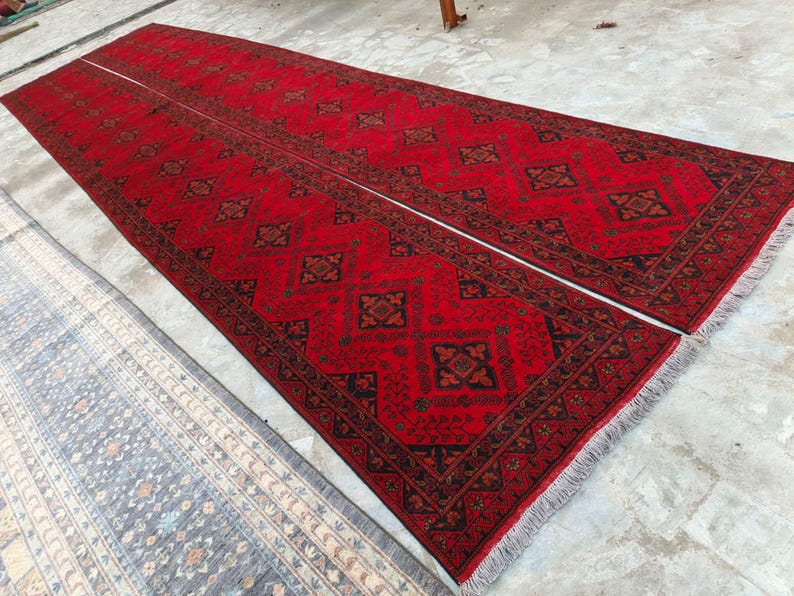 K&ouml;nnte beinhalten: Zwei lange, rechteckige rote Teppiche mit komplizierten geometrischen Mustern in Schwarz und Braun. Die Teppiche haben an den Enden Fransen und sind nebeneinander platziert. Ein kleinerer, gemusterter Teppich in Blau- und Beiget&ouml;nen ist teilweise sichtbar.