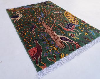 May include: Green rectangular rug with a tree and bird design. The rug features a central brown tree with colorful birds, yellow leaves, and blue flowers. The rug has a white fringe.