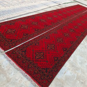 K&ouml;nnte beinhalten: Zwei lange, rechteckige rote Teppiche mit komplizierten geometrischen Mustern in Schwarz und Braun. Die Teppiche haben ein zentrales Design und einen detaillierten Rand mit Fransen. Die Teppiche sind nebeneinander platziert.
