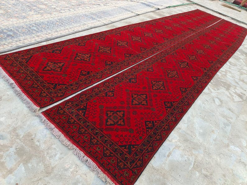 K&ouml;nnte beinhalten: Zwei lange, rechteckige rote Teppiche mit komplizierten geometrischen Mustern in Schwarz und Braun. Die Teppiche haben ein zentrales Design und einen detaillierten Rand mit Fransen. Die Teppiche sind nebeneinander platziert.